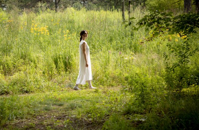 A woman with dark hair in a white robe walking pensively through a lush, green meadow.