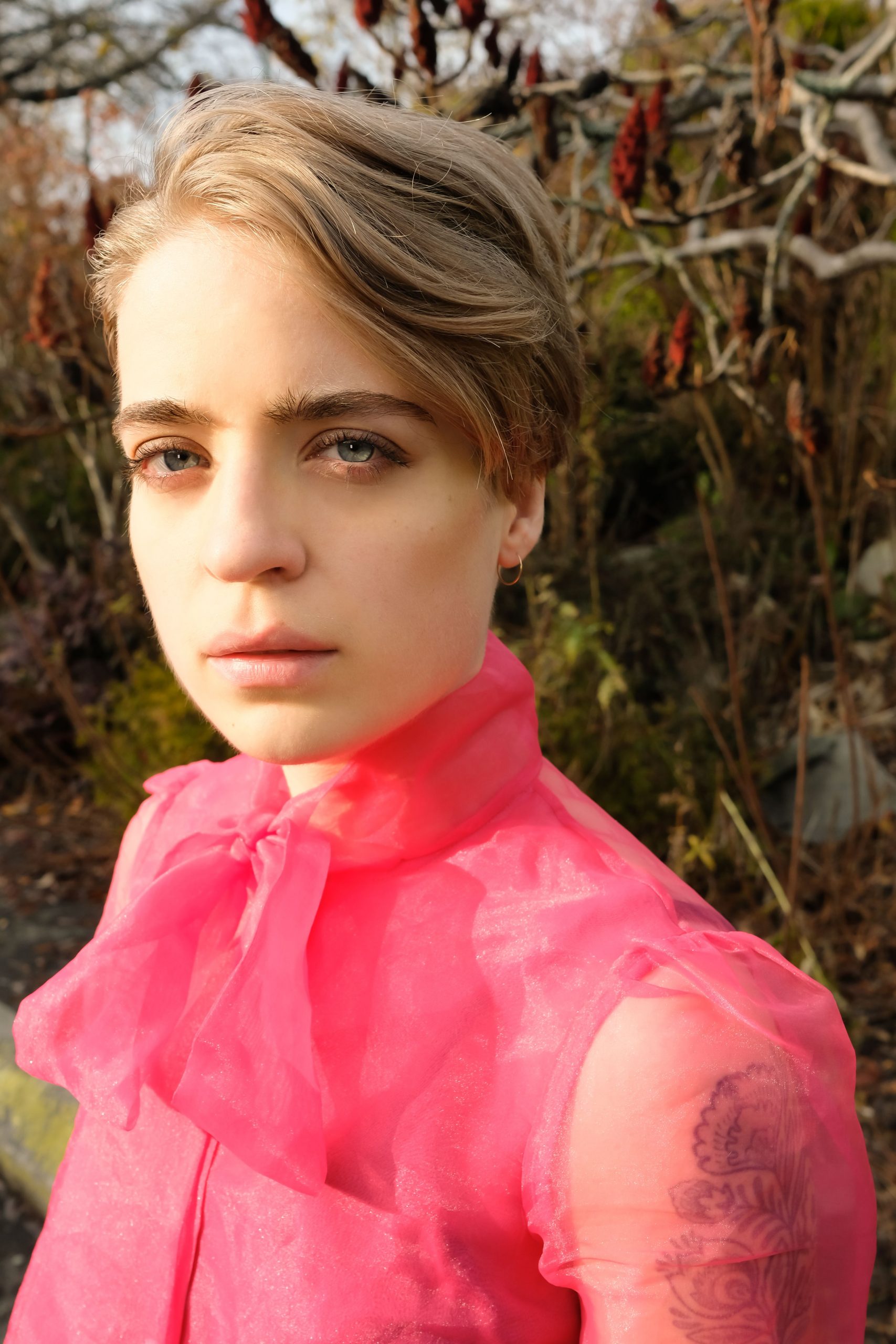 Anais West stands outdoors, pictured from the sternum up, looking into the camera. They have blond hair and blue eyes, and their face is illuminated by the sun on one side as they look intently into the camera. They stand in front of several tress with red flowers, which are just out of focus. Anais wears a sheer hot pink blouse, with capped sleeves and a bow tied around its high neck. A tattoo is just visible beneath the sleeve on Anais' left arm.