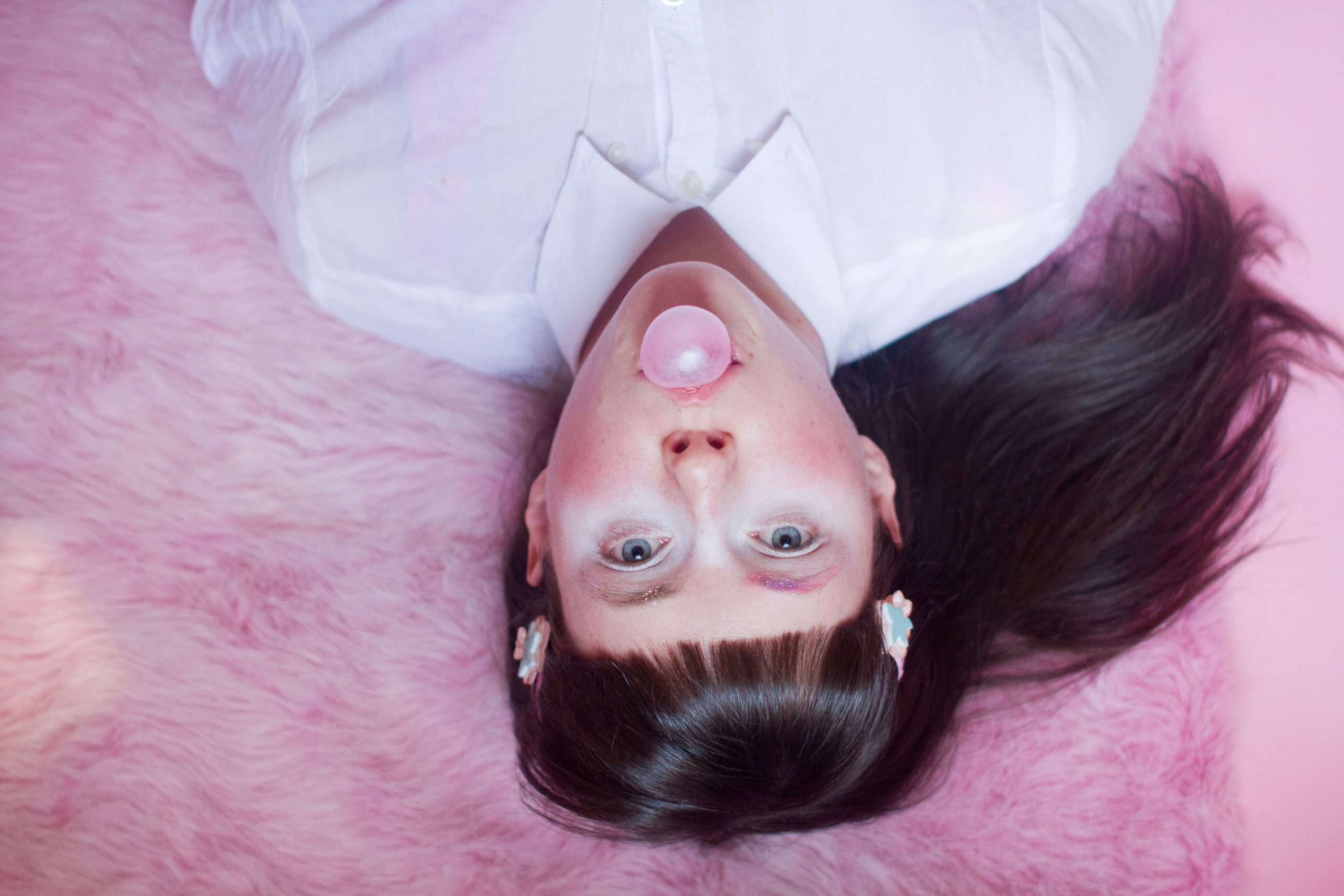 Elena Eli Belyea lays on their back in a white collared shirt, staring directly into the camera. Pictured from the sternum up, they lay on a light pink faux-fur rug, blowing bubbles with pink gum as they look back at the viewer. Their long dark hair is clipped back with light blue and pink star-shaped barettes, and swept out to their left behind them.