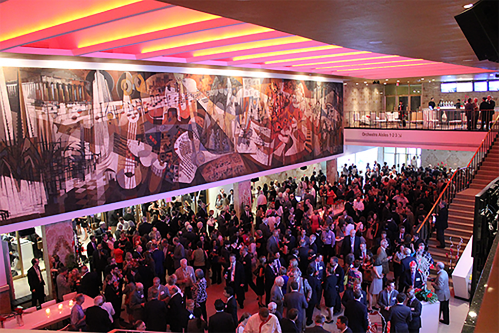 Looking down from the second floor, we can see Meridian Hall's main lobby in the middle of a networking event. Patrons are lit by pink overhead lights, which emphasise the mural by Canadian artist York Wilson.