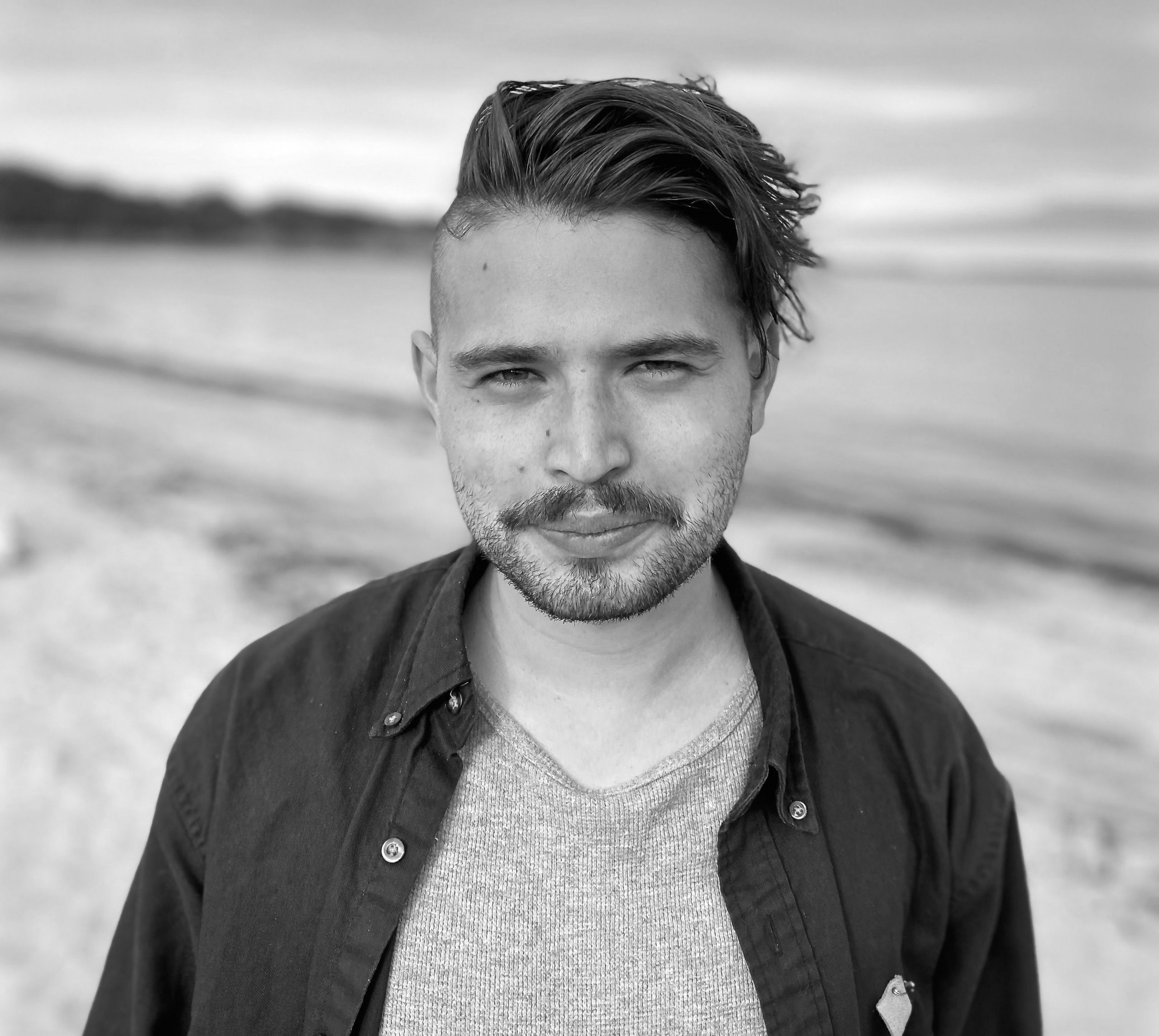 Philip Jonah Logan Geller is pictured in black and white from the sternum up. They wear a dark button-down shirt, open over a light coloured v-neck tee. The sport a half-smile as they gaze into camera, the waves of a beach visible behind them. They have dark eyes and short dark hair, trimmed on the sides, with a trimmed moustache and beard.