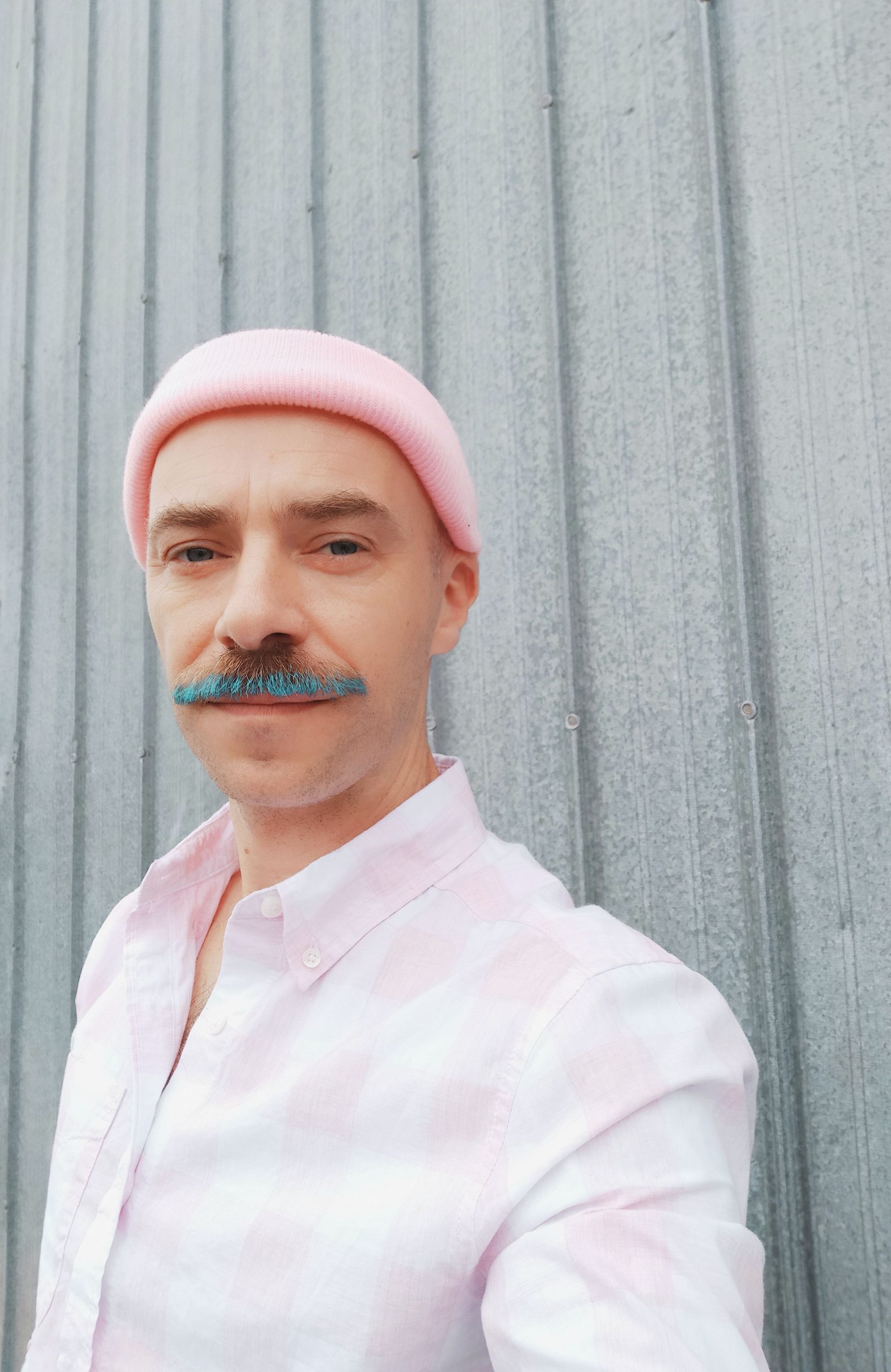 Richie Eilcox stands in front of a grey cement wall, looking directly into the camera. They are picnture from the shoulders up, wearing a pale pink button-down shirt and pink beanie. The lower part of their moustache is bright blue, and they are half-smiling as they look at the viewer.