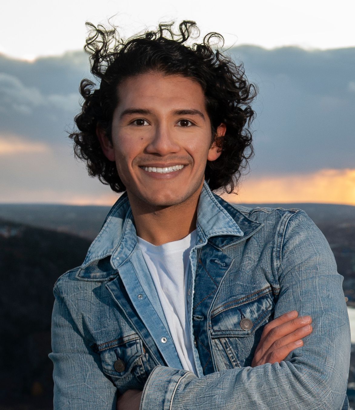 Santiago Guzmán stands outdoors in front of a sunset, pictured from the sternum up with his arms crossed. He has dark eyes and curly hair, which is blowing in the breeze. Santiago smiles widely at the camera; he wears a white t-shirt and denim jacket.