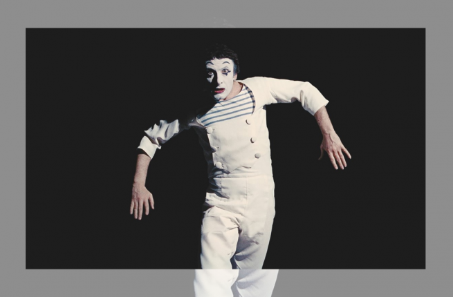 Famed French mime Marcel Marceau dresses as Bib the clown, his most famous creation. Bib wear a white one-piece jumpsuit over a black and white striped shirt, his stark white face emphasized by bold black eyebrows. his arms are outstretched, bending at the elbow, as if a marionette hanging from a string.