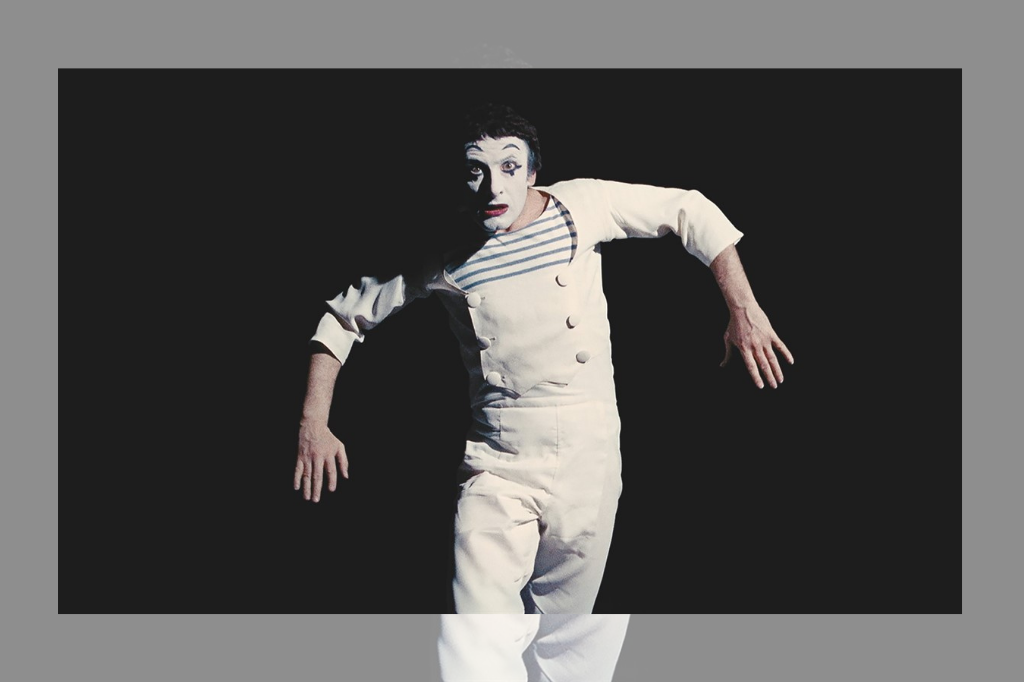 Famed French mime Marcel Marceau dresses as Bib the clown, his most famous creation. Bib wear a white one-piece jumpsuit over a black and white striped shirt, his stark white face emphasized by bold black eyebrows. his arms are outstretched, bending at the elbow, as if a marionette hanging from a string.