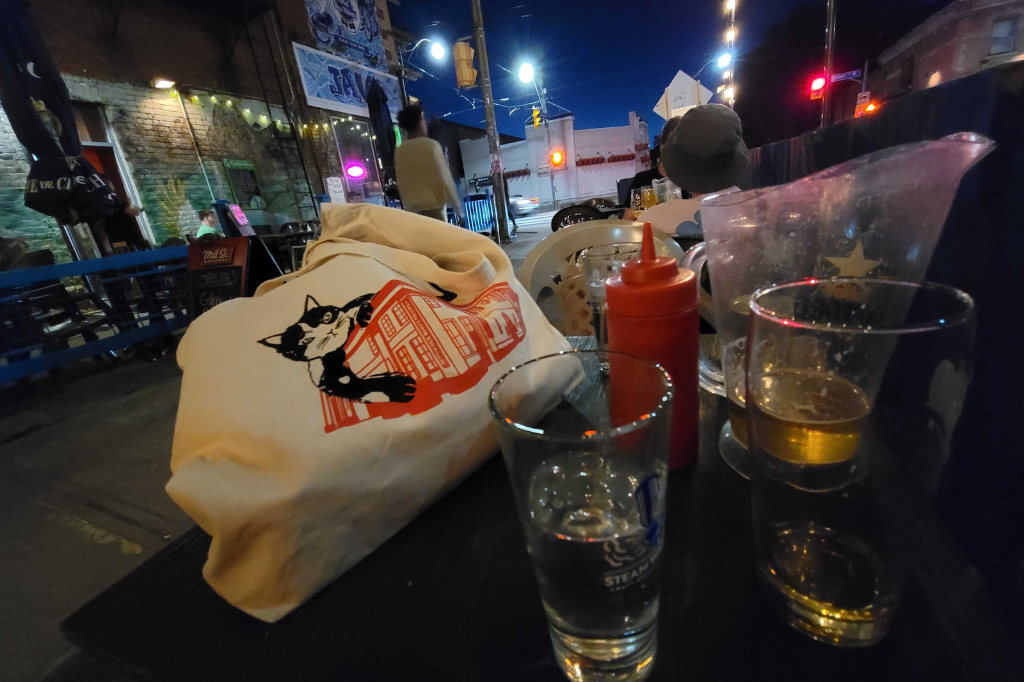 A cloth bath and ketchup bottle sit next to two half-full glasses and a pitcher of beer on a small table. A graffiti-covered wall is lit by colourful string lights, vibrant in the night sky.