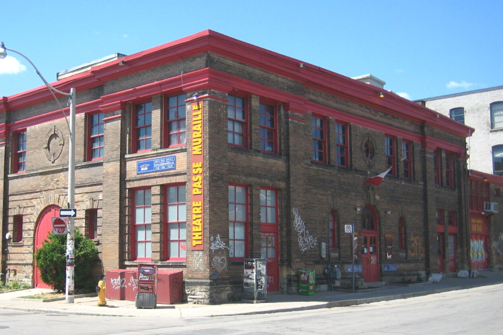 A street view of Theatre Passe Muraille, showcasing its red roof and brick exterior placed against a bright blue sky.