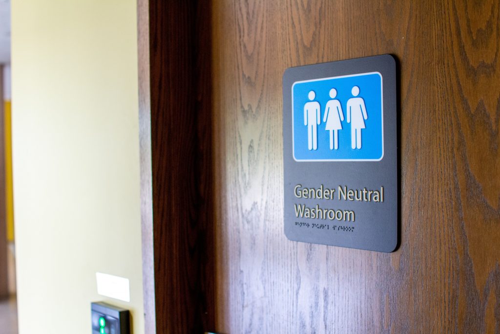 A close-up image of the door to one of Stratford Festival's gender neutral washrooms. A black sign on the wooden door features a blue washroom logo with three white figures. The sign reads "gender neutral washroom, and includes a braille translation.