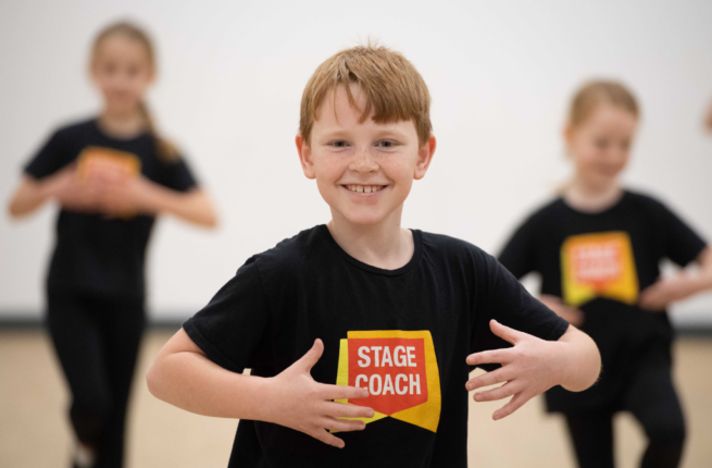 Three small children hold dance poses, smiling at the camera. they each wear black t-shirts emblazoned with the Stagecoach Performing Arts logo.