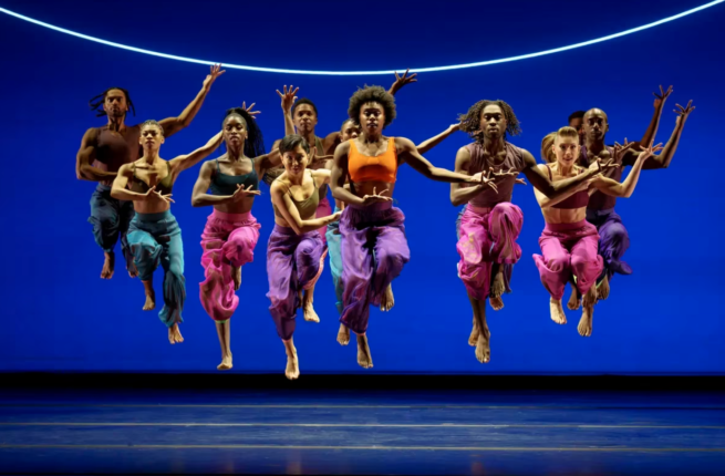 Several dancers wearing brightly coloured tank tops and loose pants leap as one into the air, onstage with a dark blue background.