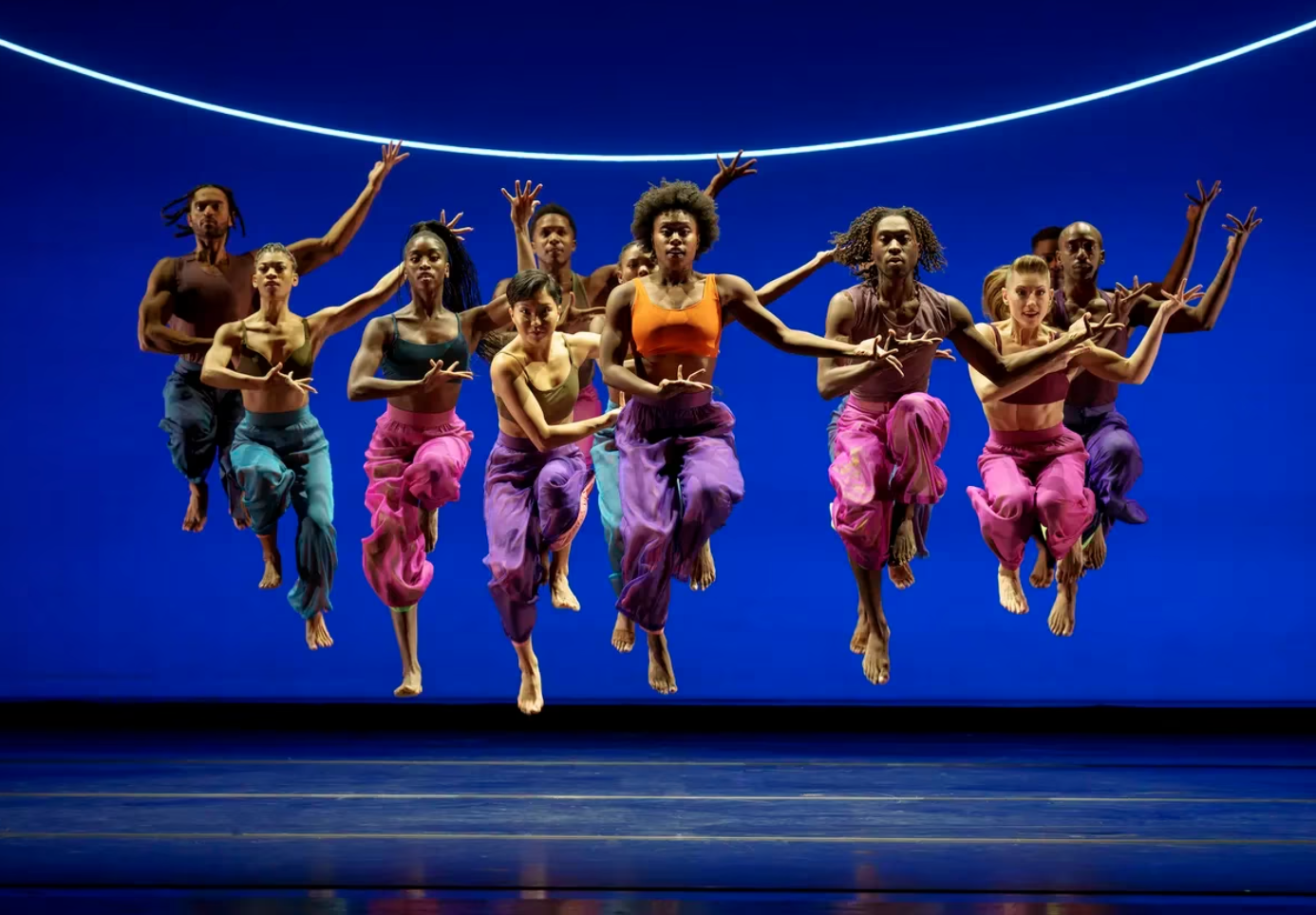 Several dancers wearing brightly coloured tank tops and loose pants leap as one into the air, onstage with a dark blue background.
