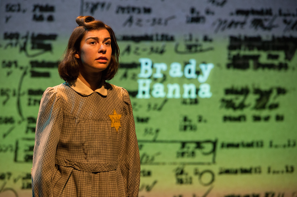 A production image from YPT's Hana's Suitcase. A young girl in a torn green dress is mid-speech. There is a yellow star with the word "Jude" stitched into it on her chest. Set & Costume Design by Teresa Przybylski; Lighting Design by Andrea Lundy; Photo by Cylla von Tiedemann.