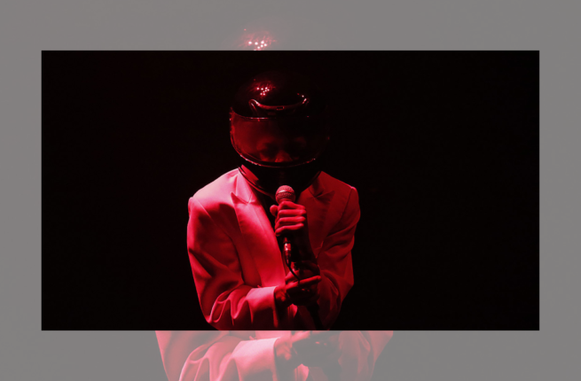 Person standing on stage in suit jacket, wearing a helmet with a visor and speaking into a microphone they are holding. Bathed in red light with a black background.