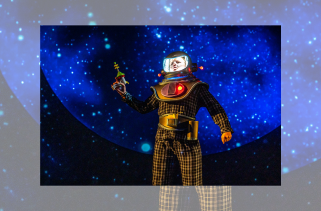 A man in a yellow and black plaid astronaut suit stands on stage in front of a backdrop of outerspace.