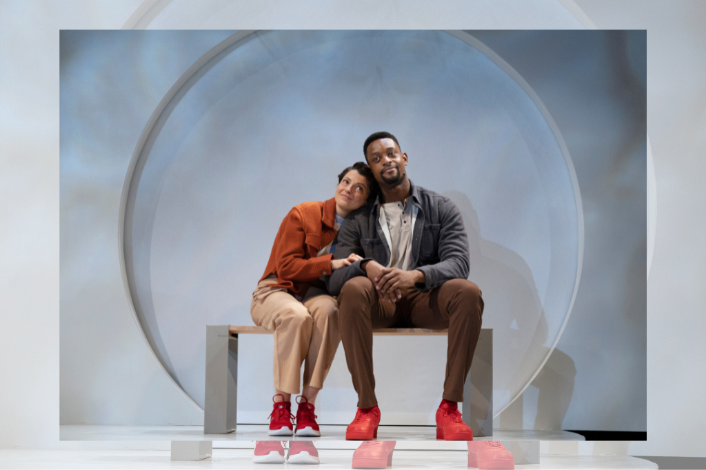 A woman and a man sit on a bench on stage, with a grey background. The woman wears a red jacket and red shoes, and leans onto the man, clutching his arm. They are both smiling upwards gently.