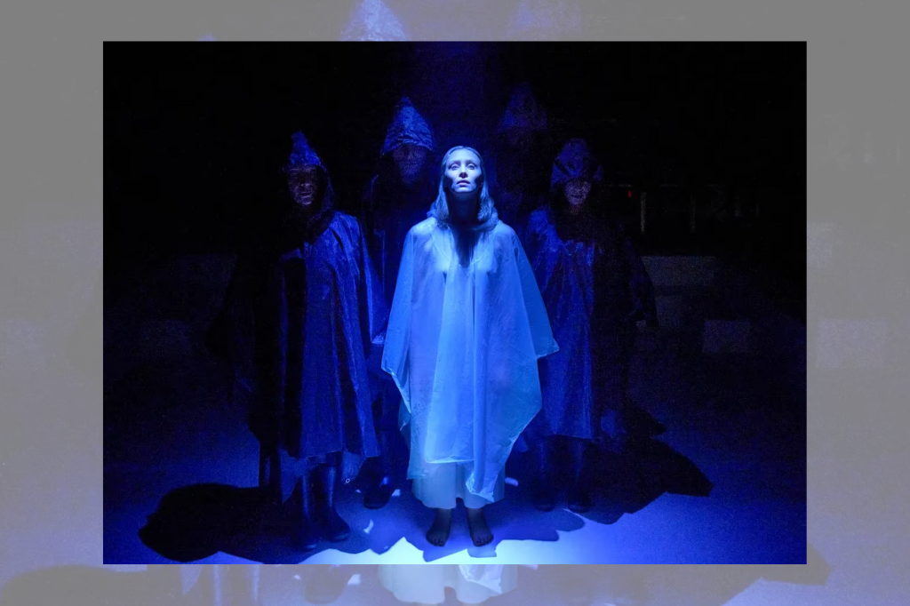 A woman stands in a sheer, white dress with a hood in a blue light on stage, surrounded by three dark figures in black hooded cloaks.