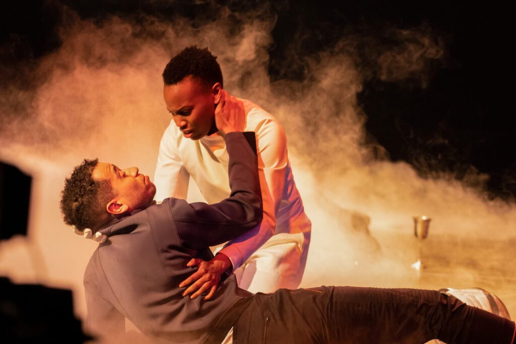 From left: Austin Eckert and Amaka Umeh in Hamlet. Umeh holds Eckert as he falls back, reaching up towards her face. Smoke billows behind the two figures, illuminated on an otherwise dark stage. Photo by Jordy Clark. Image courtesy of the Stratford Festival.