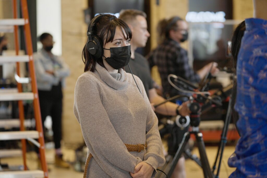 Genna Dixon during filming of Northern Tracks in December 2022. She stands with her hands clasped in front of her. She wears a headset and a mask, and stands in front of several members of the crew, ready to film. Image courtesy of Stratford-Festival.