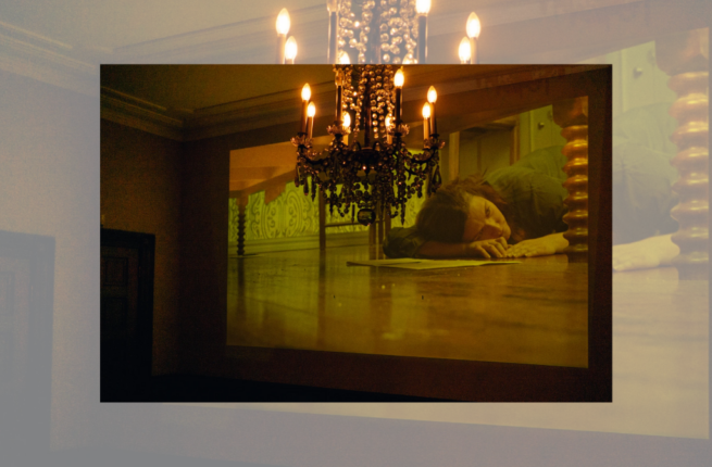 Chandelier in foreground with a projection on background wall of a woman lying on the floor beneath a table, looking lost in thought.