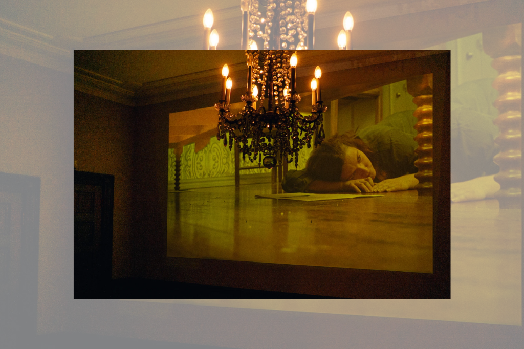 Chandelier in foreground with a projection on background wall of a woman lying on the floor beneath a table, looking lost in thought.
