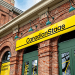 A view of the front of Canadian Stage's exterior, taken from an angle. A large, red-brick building with green doors and windows. A sign reading "canadian stage" hangs above the front doors, with green lights above to illuminate it.
