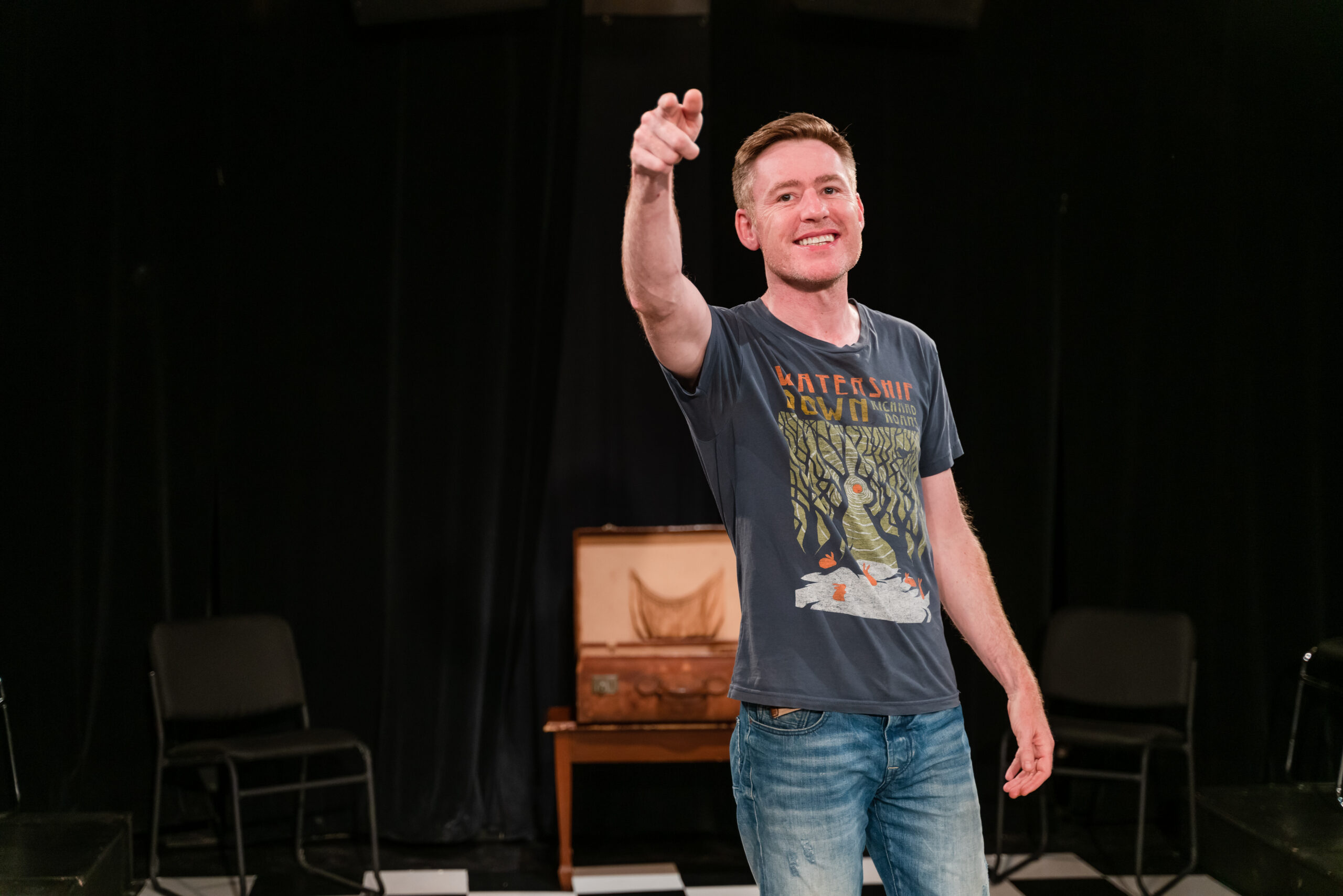 Gavin Crawford stands on a stage, pointing out into the audience. A single, empty chair sits behind him. Crawford wears a grey graphic t-shirt and jeans, and smiles as he speaks.