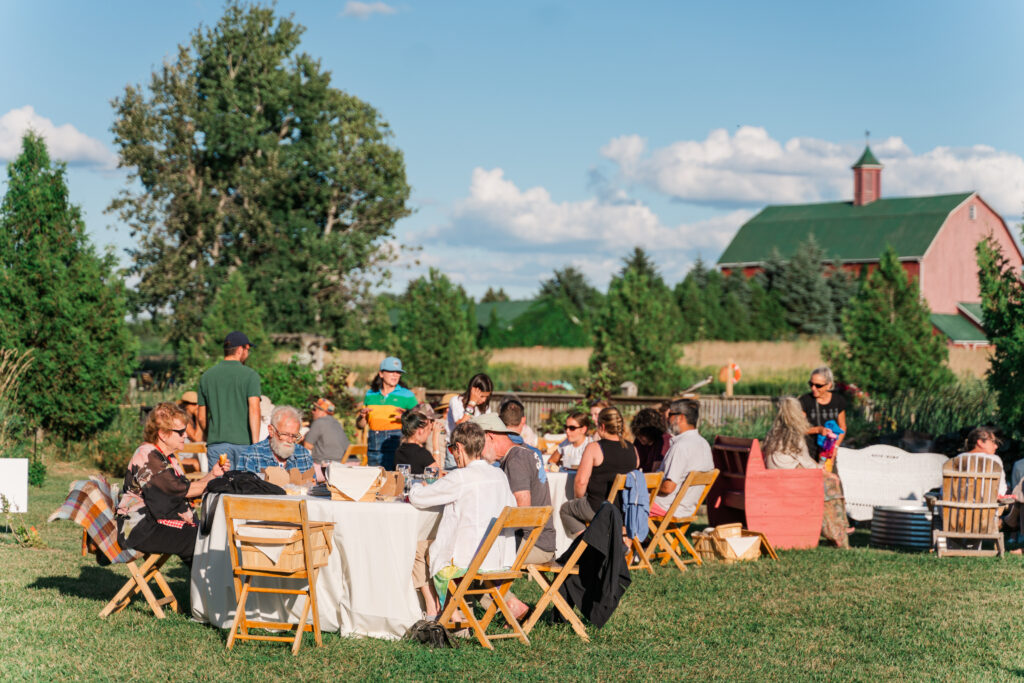 Pre-show picnics at The Eddie Hotel & Farm before Flight Festival of Contemporary Dance (2022). People sit at round tables covered with white tablecloths, eating from picnic baskets. It's a sunny day, and the green roofed barn is visible in the background.