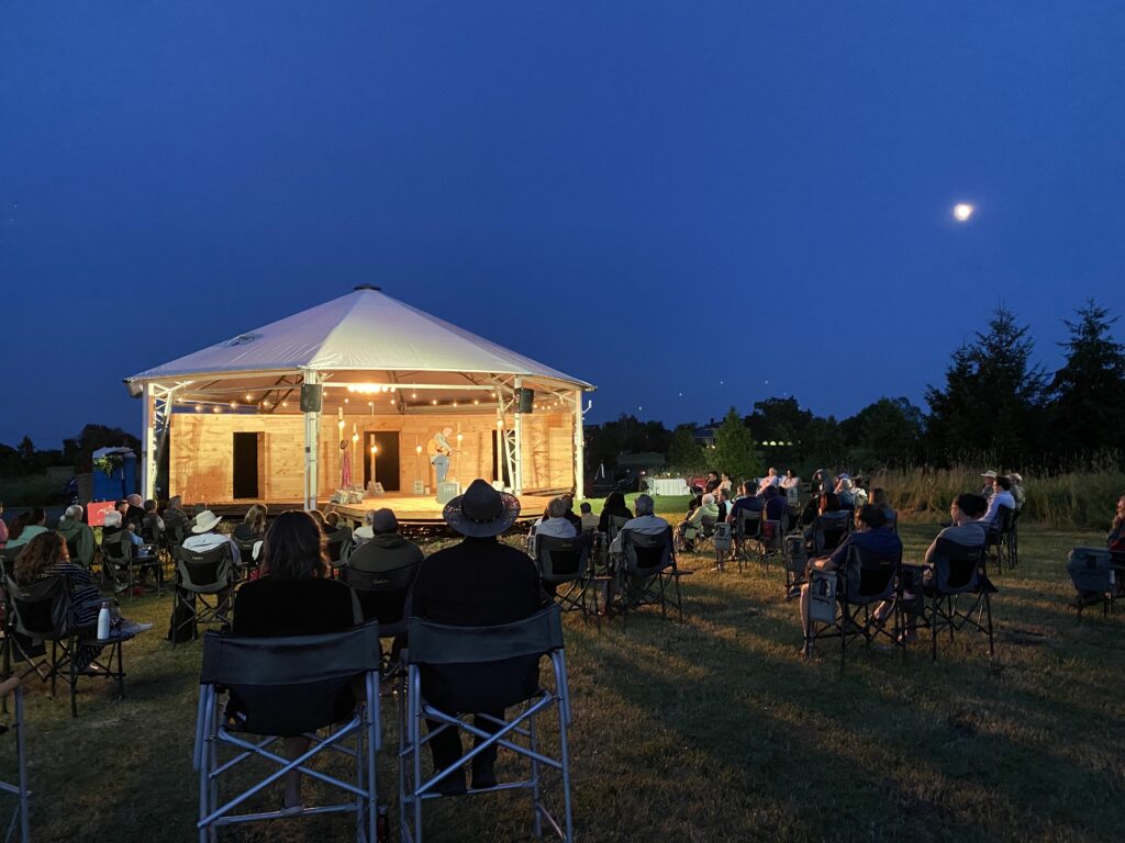 A nighttime view of the Eddie Pavilion. Audiences sit in folding chairs, watching Beau Dixon performing Springhill (2022). The pavilion is lit with soft string lights, illuminating the white structure against a deep blue evening sky.