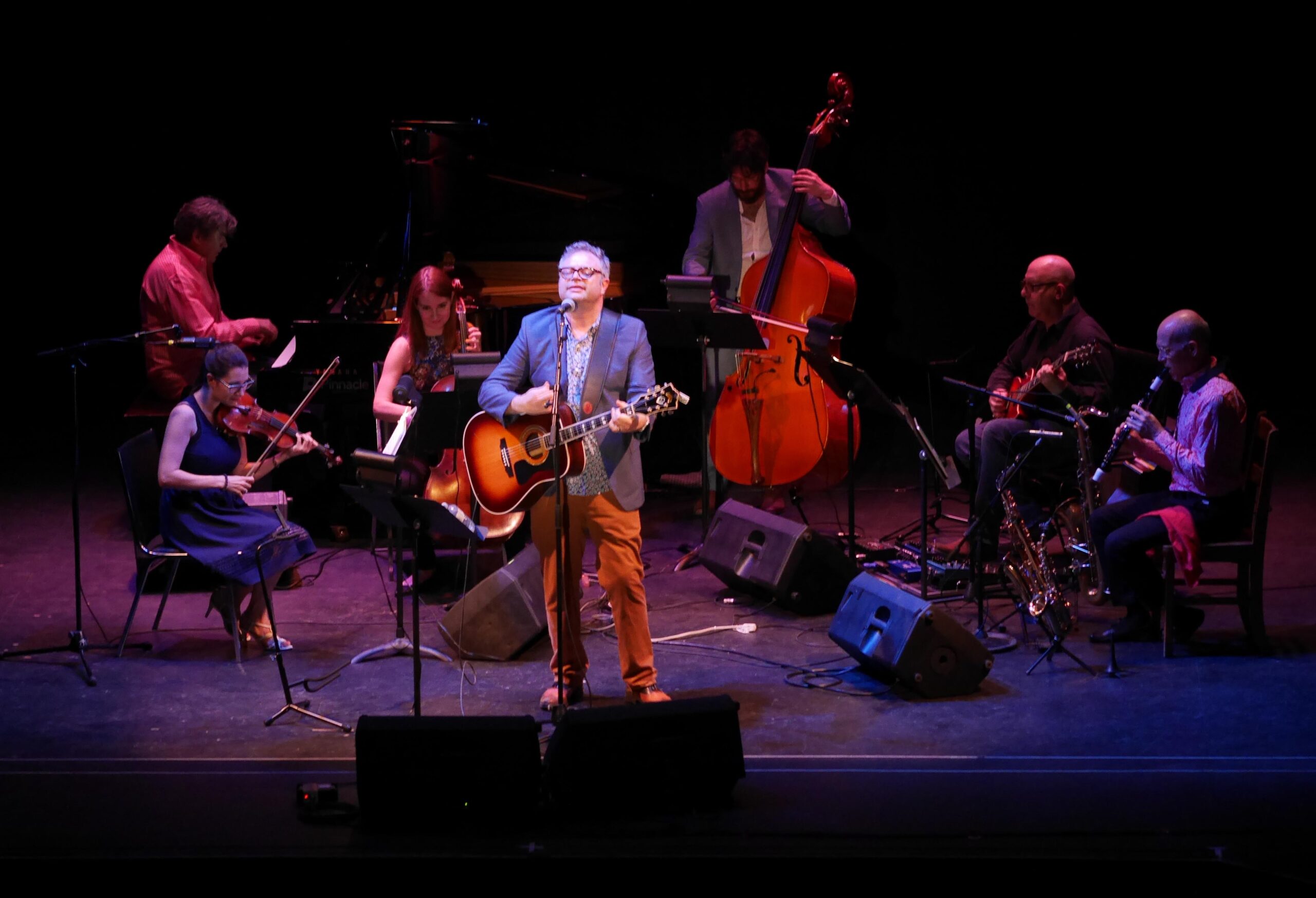 Stephen Page stands at a microphone, guitar in hand, as he sings. He is surrounded by a six-piece musical ensemble of string instruments, all faintly lit with red and blue lights. Image provided by County Stage Company.