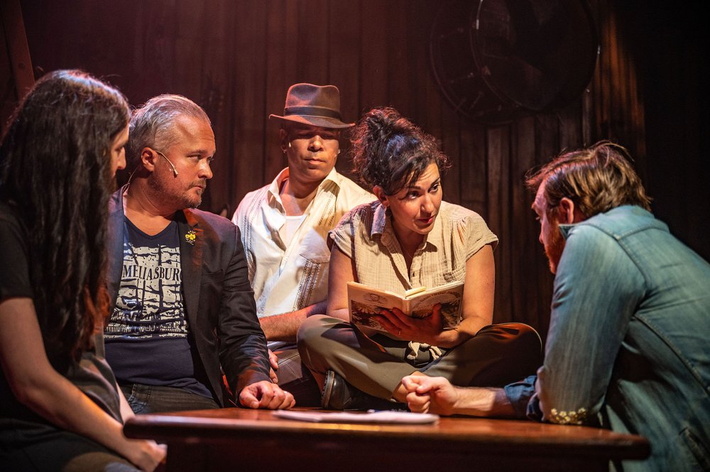 (left to right) Hailey Gillis, Andrew Penner, Beau Dixon, Raha Javanfar, and Frank cox-O'Connell in World Premiere of The Shape of Home, performed at The Eddie Red Barn Theatre as part of County Stage Company's 2023 Festival. The five musicians crowd around a wooden table, which Javanfar is seated on, looking at the book in her hands. Image by Dahlia Katz.