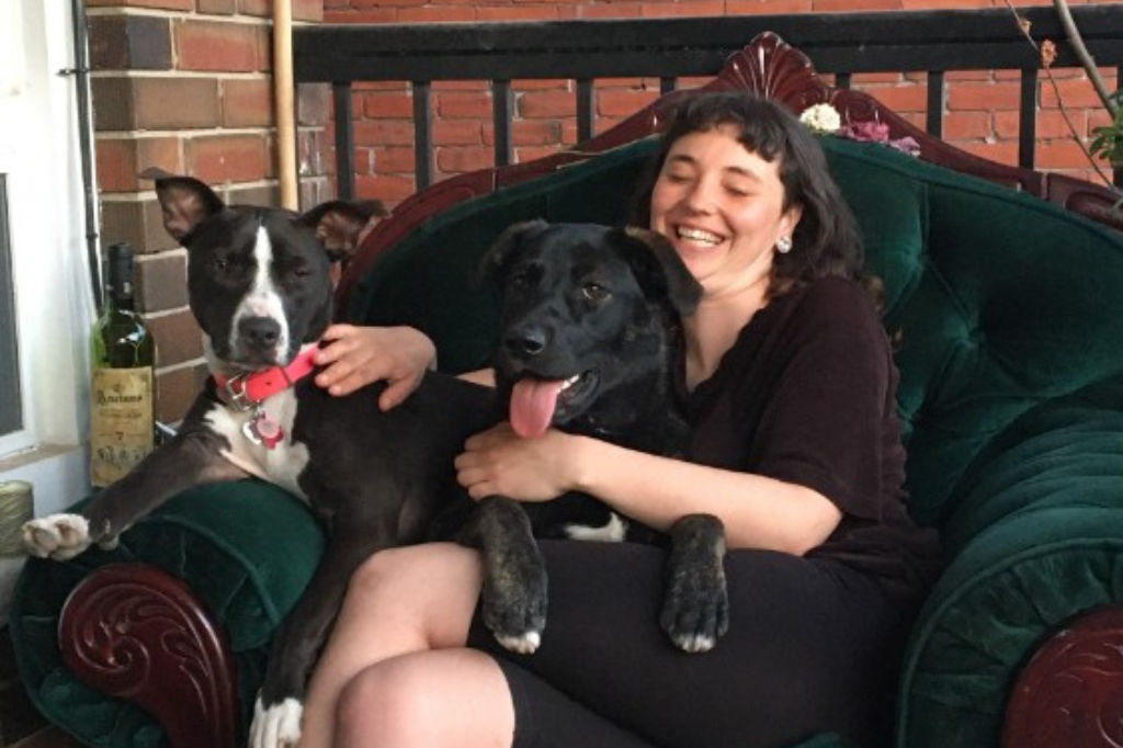 Heather sits on a dark green, velvet love seat, with two dogs in her lap. She wears dark clothing, and laughs as she pets the pups. Her dog, Lola, a black and qhite muscle-breed cross with a pink collar squints at the camera, while Stevie, a black dog, smiles with their tongue out.