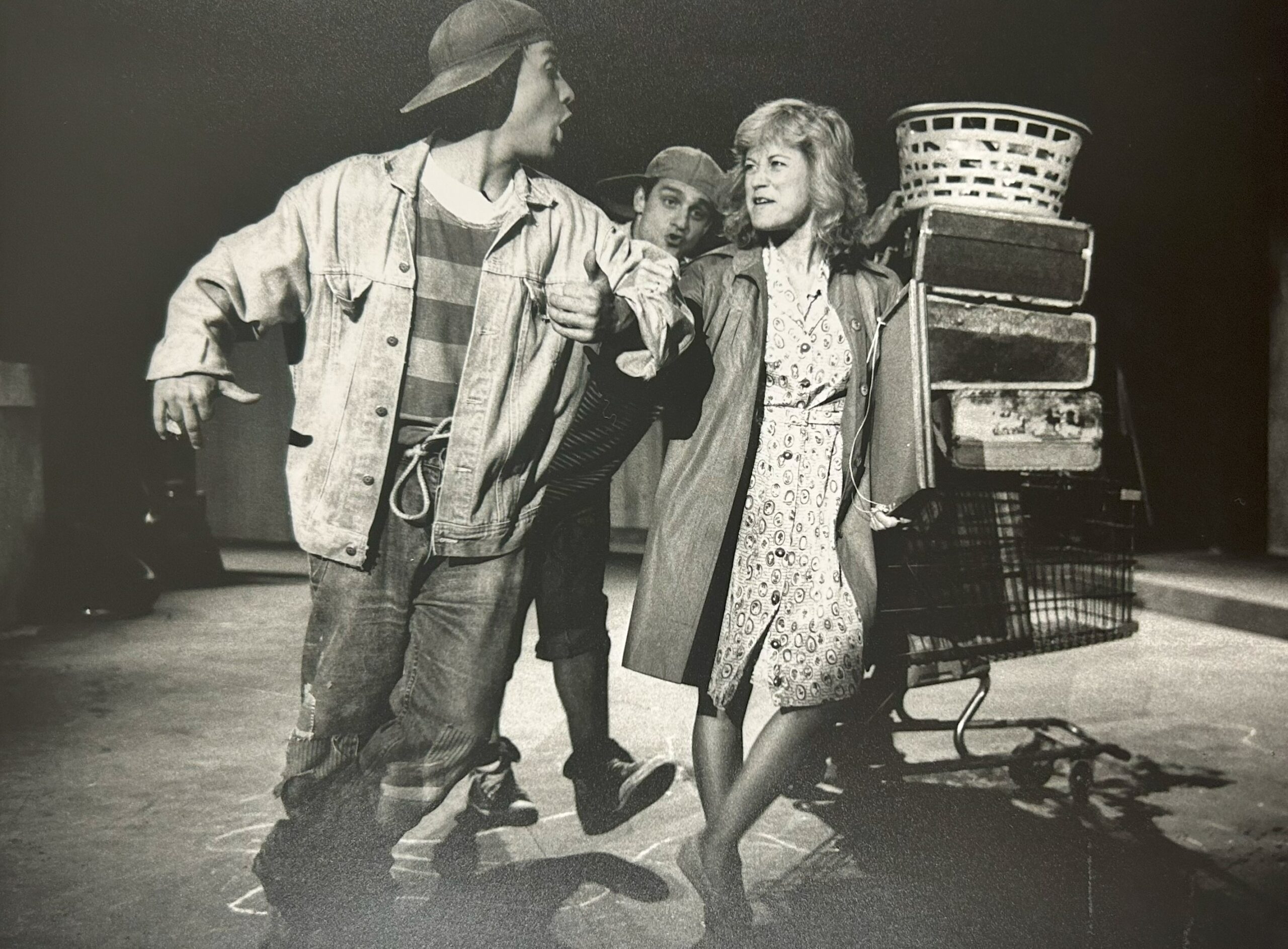 A black-and-white photograph depicting three performers in the 1988 Dream in High Park production of Blood Brothers. The two men wear baseball caps, jeans and jackets while the woman sports a patterned dress and cardigan.