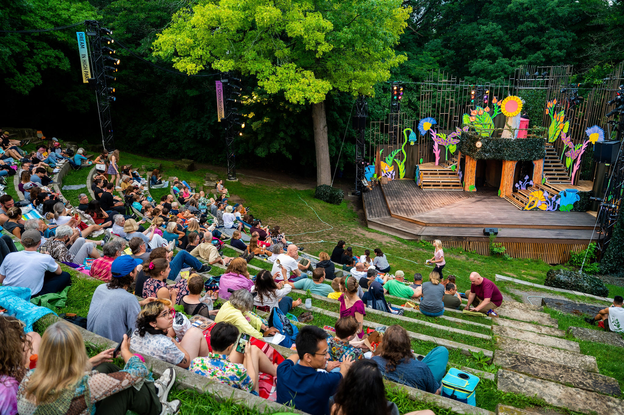 A full audience waits to watch the summer production of As You Like It in Dream in High Park's 2022 programming. The stage is covered with large, colourful flowers. Image courtesy of Canadian Stage.