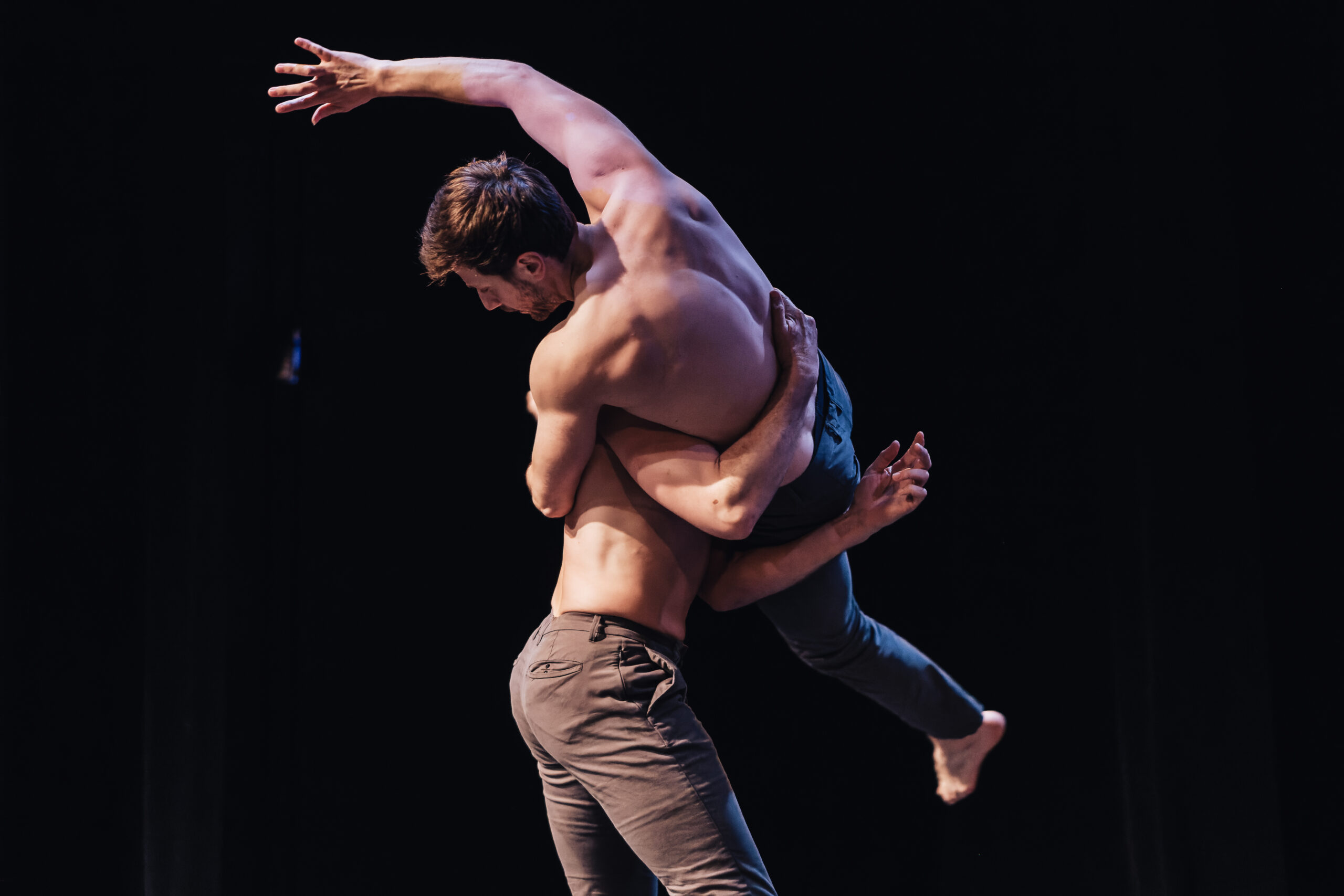 Two men perform on stage, one lifting the other in his arms as he twists in an acrobatic position. Both men are shirtless and wear neutral-coloured trousers. Original image by Curtis Perry.