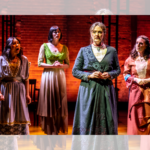 4 women in dresses stand on stage with a red brick background. They sing and look distressed.