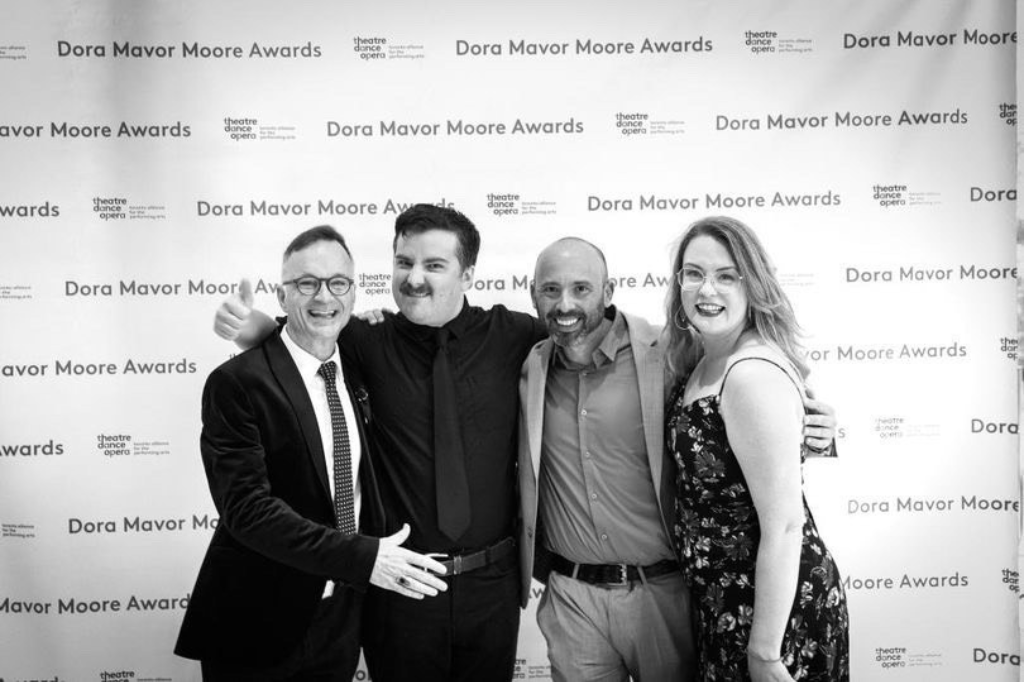 Jonathan Wilson, Curtis Campbell, Daniel Krolik and Rachel Kennedy pose in front of the photo backdrop at the 2023 Dora Mavor Moore Awards. They are dressed for a formal occasion: Wilson, Krolik, and Kennedy smile at the camera, while Campbell gives a "thumbs up" with a grimace.