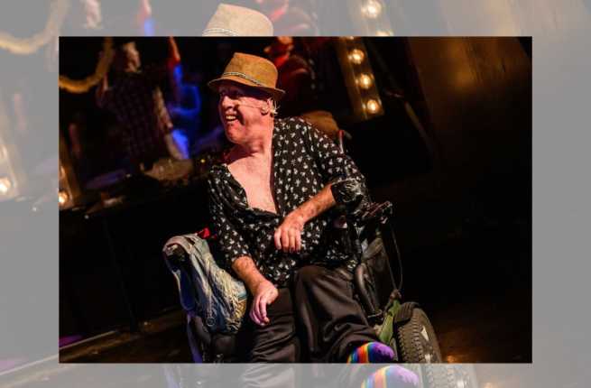 A man wearing a fedora, unbuttoned shirt, and rainbow socks sits smiling onstage in a wheelchair.
