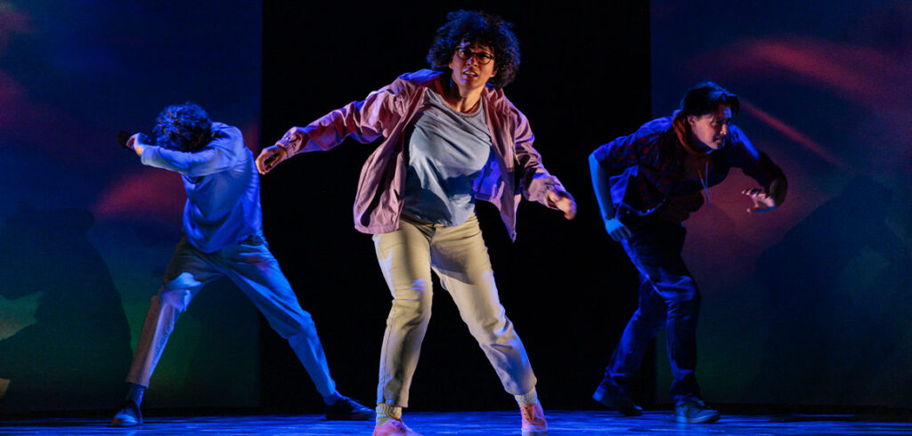 From left: Noah Beemer, Celeste Catena, and Robert Markus perform in A Wrinkle in Time at the Stratford Festival. Catena wears a pink jacket and tan pants, while the two men wear t-shirts and trousers. The stage is lit with blue light, and the three seem off-balance, as if floating. Photo by David Hou.
