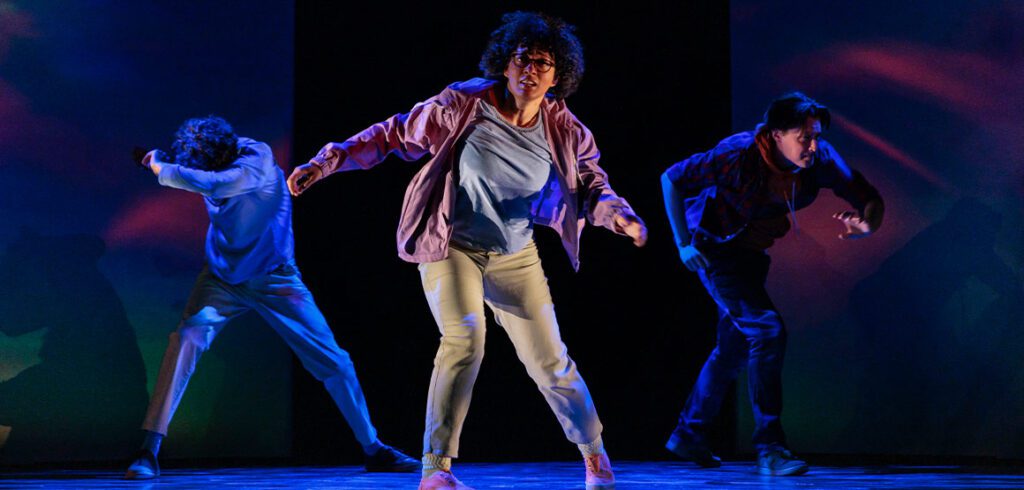 From left: Noah Beemer, Celeste Catena, and Robert Markus perform in A Wrinkle in Time at the Stratford Festival. Catena wears a pink jacket and tan pants, while the two men wear t-shirts and trousers. The stage is lit with blue light, and the three seem off-balance, as if floating. Photo by David Hou.