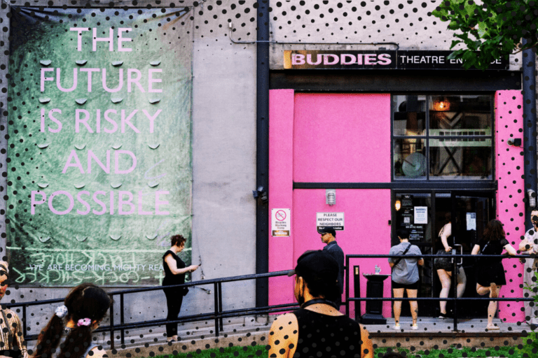 The exterior of Buddies in Bad Times Theatre. The bubblegum-pink double doors are open, allowing a crowd of visitors to enter. Above the door is the company's logo, also in pink. To the left of the door, a large green banner hangs against a concrete wall. Pale pink text on the banner reads 