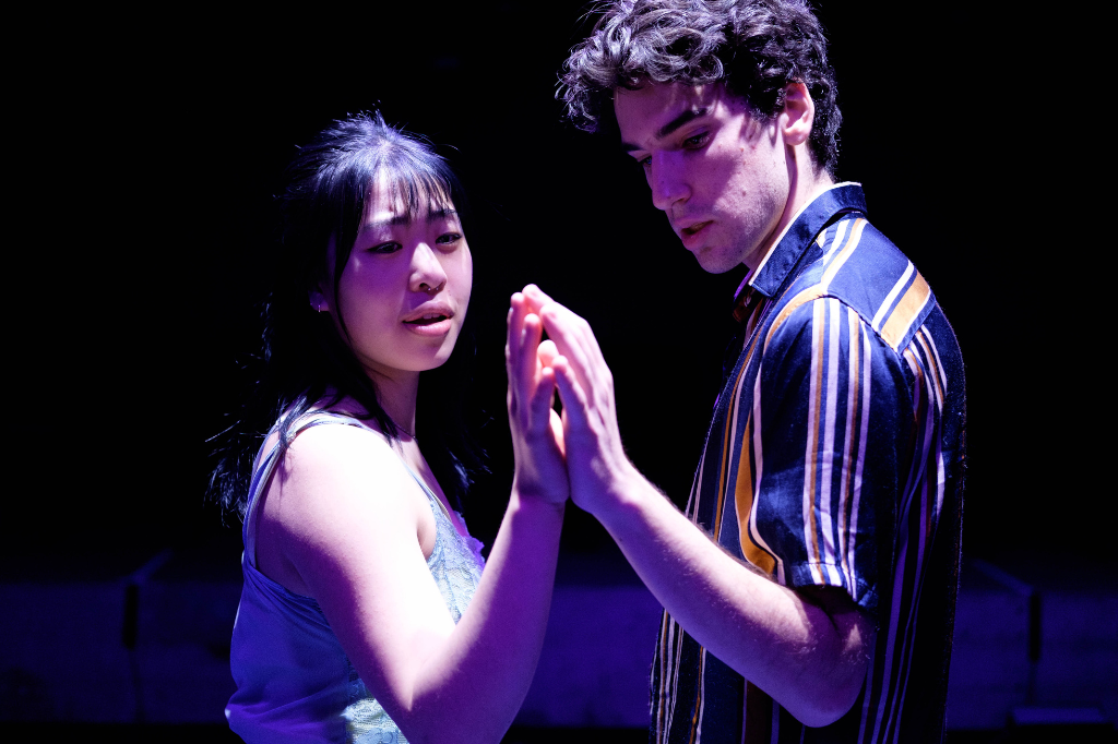 Production photo from Next Stage Festival production "ECHO." Echo and Narcissus smile as they press their hands against each others, glowing in light from above.