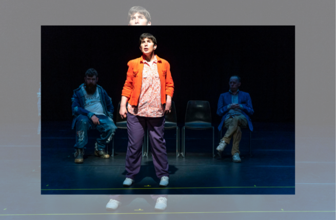Production photo of Back to Back Theatre's The Shadow Whose Prey the Hunter Becomes. A woman in an orange jacket stands centre stage; behind her, two men sit in chairs.