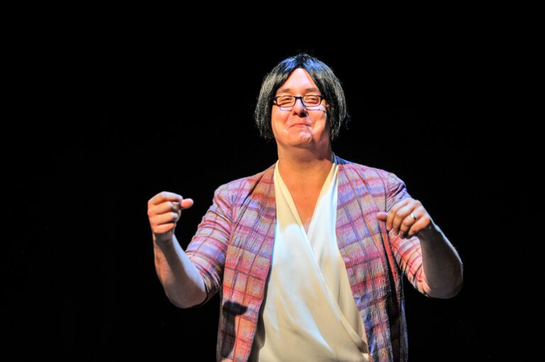 Comedian Mike Delamont wears a short grey bob-cut wig, a pink robe, and a white shirt in 