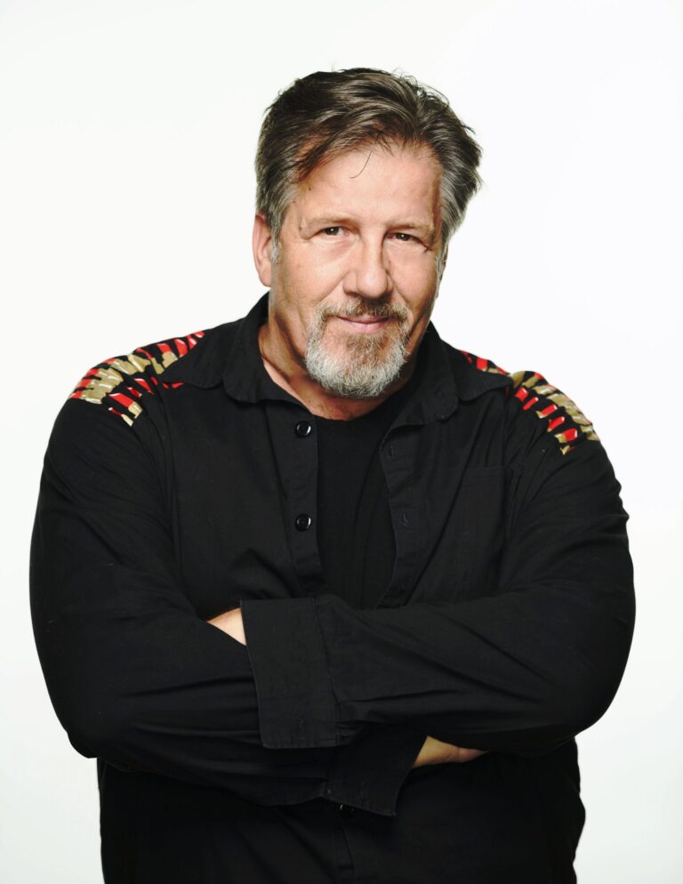 Darrin is pictured from the waist up, standing in front of a white background - he smiles casually with his arms crossed in front of his sternum. Darrin has brown hair and a silver-grey beard; he wears a black button-down black shirt, partially unbuttoned over a black t-shirt. There are red, gold and white details printed on the shoulders of his shirt - tubes of lipstick pointing in various directions.