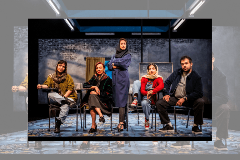 The cast of English, lined up onstage. Four of the individuals sit at school desks, gazing at the camera: some smile, others look inquisitive. The man wears a dark jacket and trousers, while the women wear brighter colours and patterned hijabs. A woman in blue with a dark hijab stands in the centre of the line, her arms crossed, staring intently into the camera with a gentle smile.
