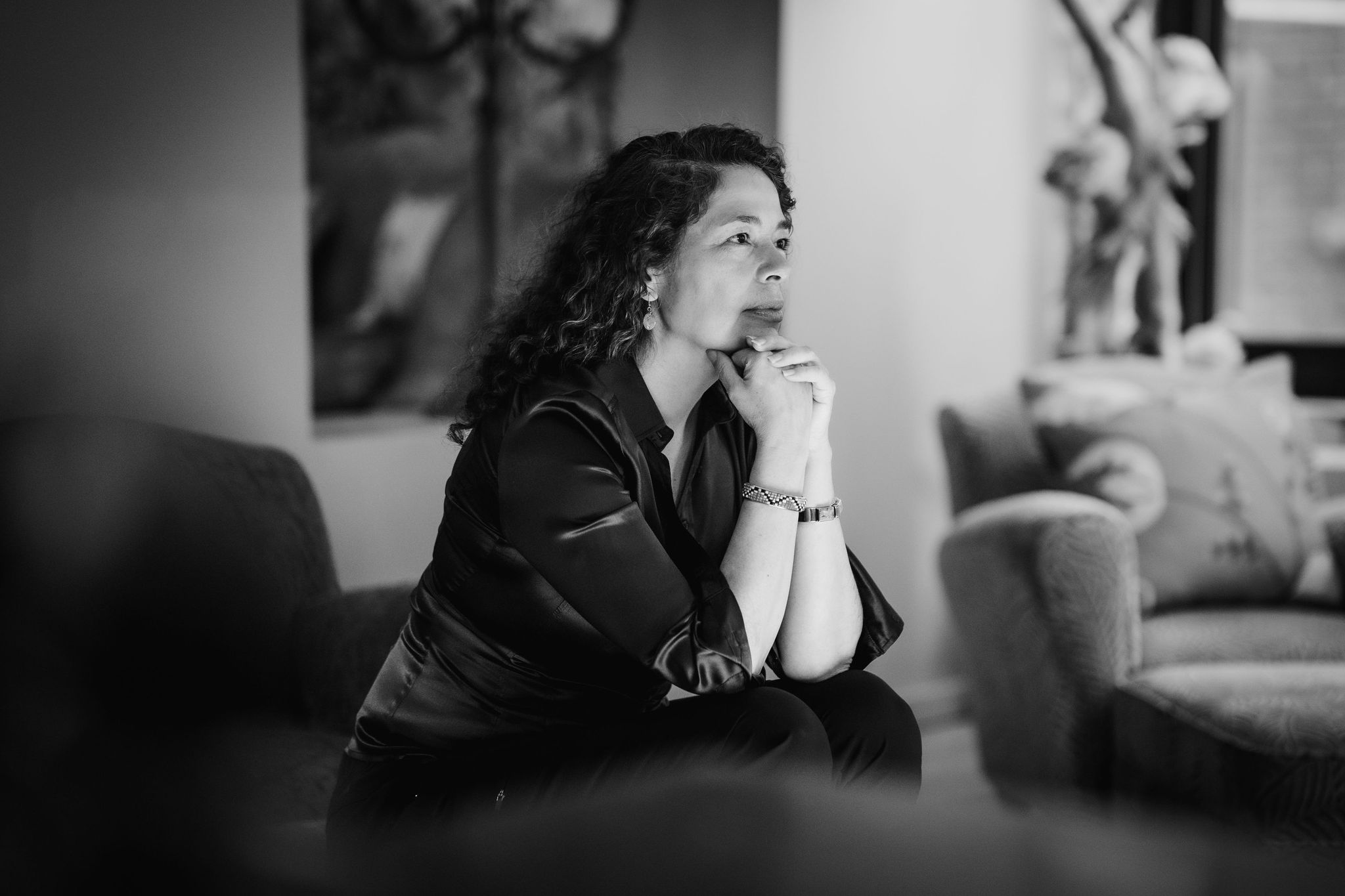 Sandra Laronde sits on a couch with art behind her, her hands drawn together in fists at her chin, looking out to the right. Black and white photo.