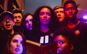 Eight artists are gathered together, their faces lit by red and blue lights, staring forward and up with a variety of expressions - some are concerned, others seemingly excited - but they are all staring intently at what they're looking at. The woman in the centre of the group is holding an open book.