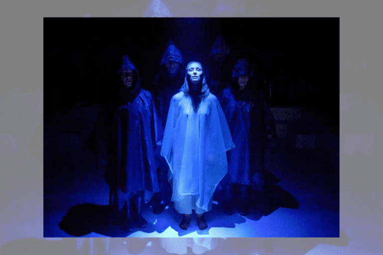 A woman stands in a sheer, white dress with a hood in a blue light on stage, surrounded by three dark figures in black hooded cloaks.