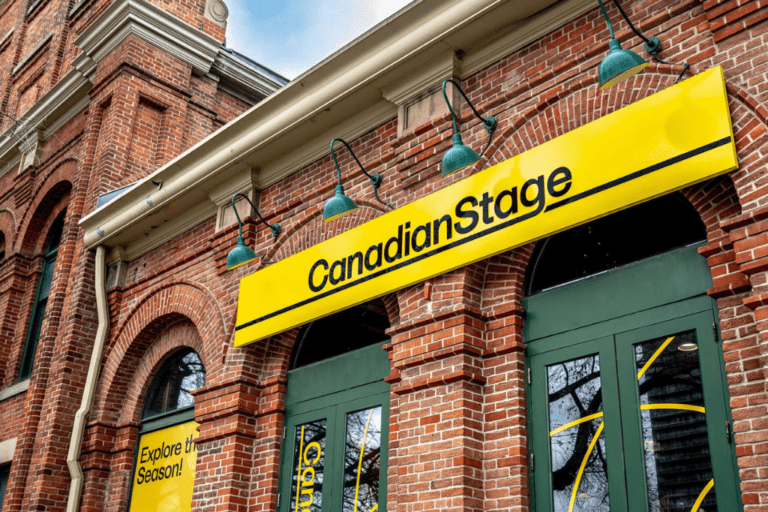A view of the front of Canadian Stage's exterior, taken from an angle. A large, red-brick building with green doors and windows. A sign reading 