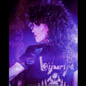 Bijuriya stands profile in purple hues with vibrant make up and a wild wig. She dawns a leather vest with her name “Bijuriya” in rhinestones.